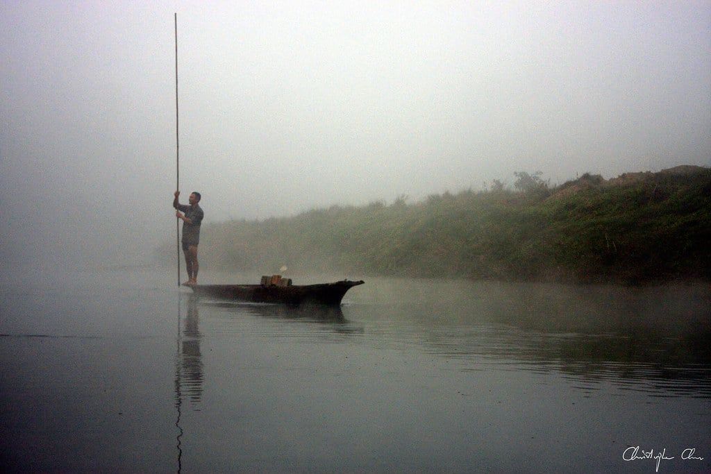 A boatman stands with a long pole in a traditional dugout canoe on the misty Rapti River in Chitwan, Nepal.